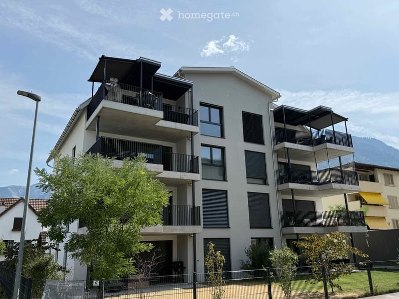 Modern multi-story residential building with balconies, large windows, and contemporary design, surrounded by greenery and a fence.