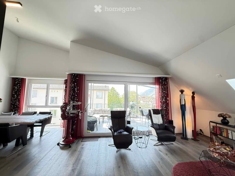 Bright living room with wooden flooring, large windows, and a balcony. Features a sofa, chairs, a table, a fireplace, and decorative statues.