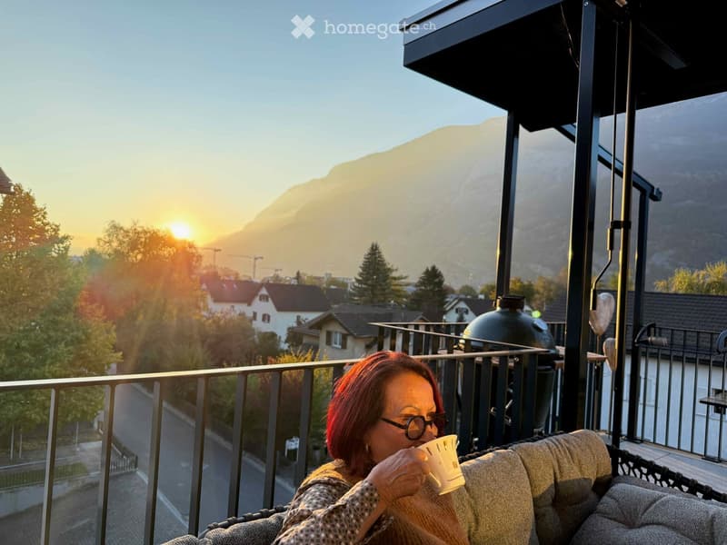 A woman is sitting on a balcony with a cup of coffee, enjoying a view of a town and mountains at sunset.