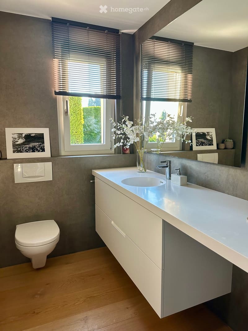 Modern bathroom with a white sink, chrome faucet, toilet, large mirror, and decorative vase with white flowers on the counter. Grey walls and wooden flooring.