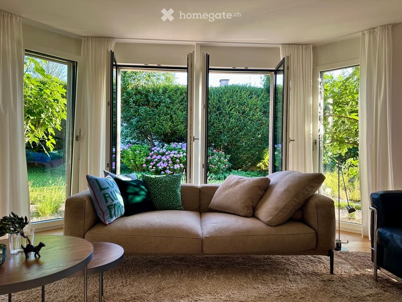 Modern living room with large windows and sliding glass doors opening to a garden, featuring a beige sofa with colorful pillows, a wooden coffee table, a side table, a floor lamp, and a black leather armchair.