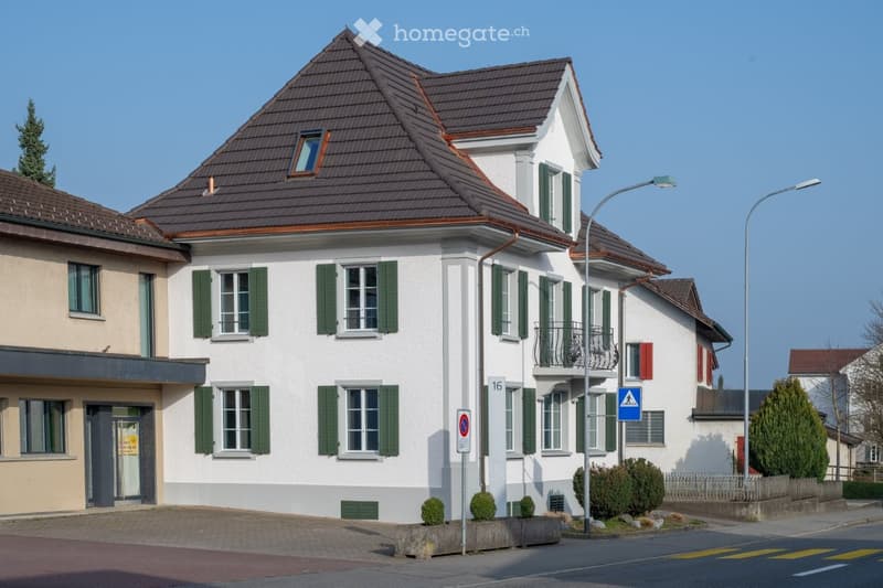 Two-story residential house with white walls, green shutters, and a dark brown tiled roof. Features include a balcony on the upper floor, small garden area, and street signage.