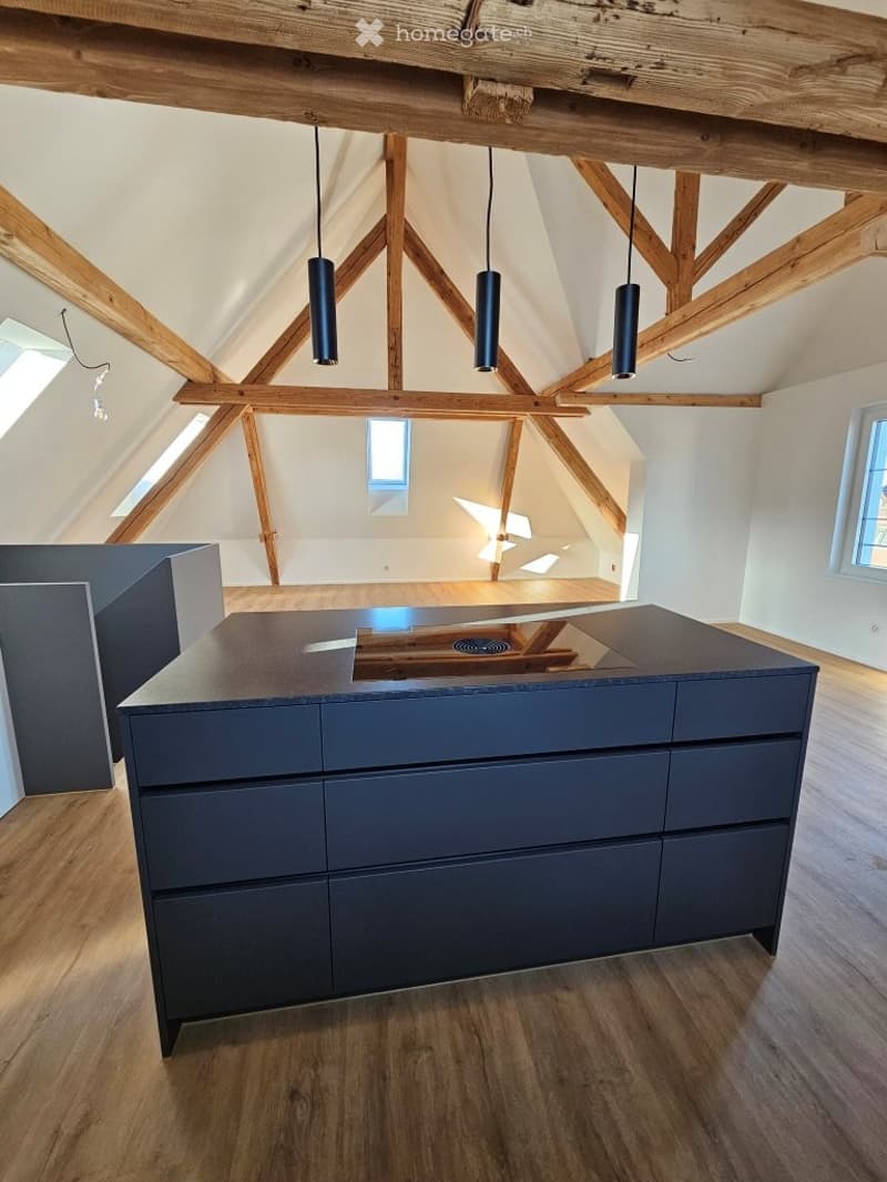 Modern kitchen with a dark grey island, wooden beams, and pendant lights. The space features exposed wooden beams and skylights, creating a bright and airy atmosphere.