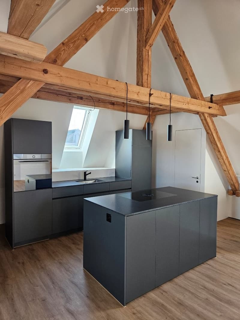 Modern kitchen with dark grey cabinets, central island, pendant lights, and exposed wooden beams on the ceiling.
