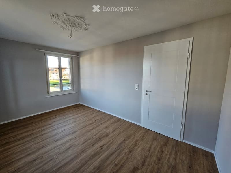 Empty room with wooden flooring, white walls, a white door, and a window showing a view of the outside.