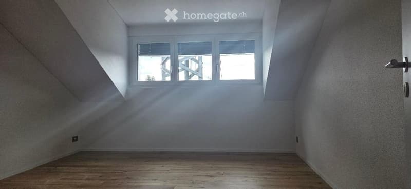 Empty attic room with wooden flooring, white walls, and a large window letting in natural light.