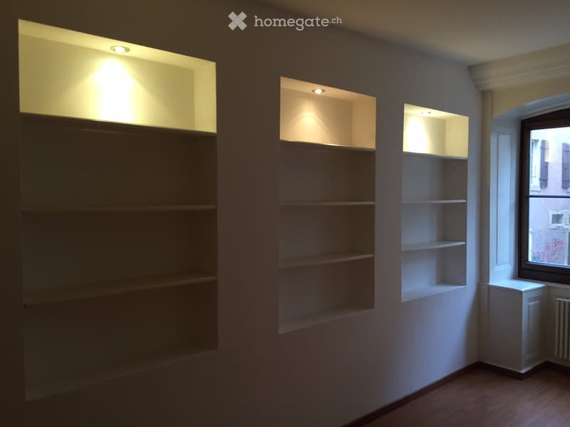 Empty room with built-in shelves and recessed lighting, wooden floor, and a window showing an exterior view.