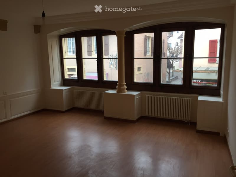 Empty room with wooden flooring, large windows, radiators, and a central column.