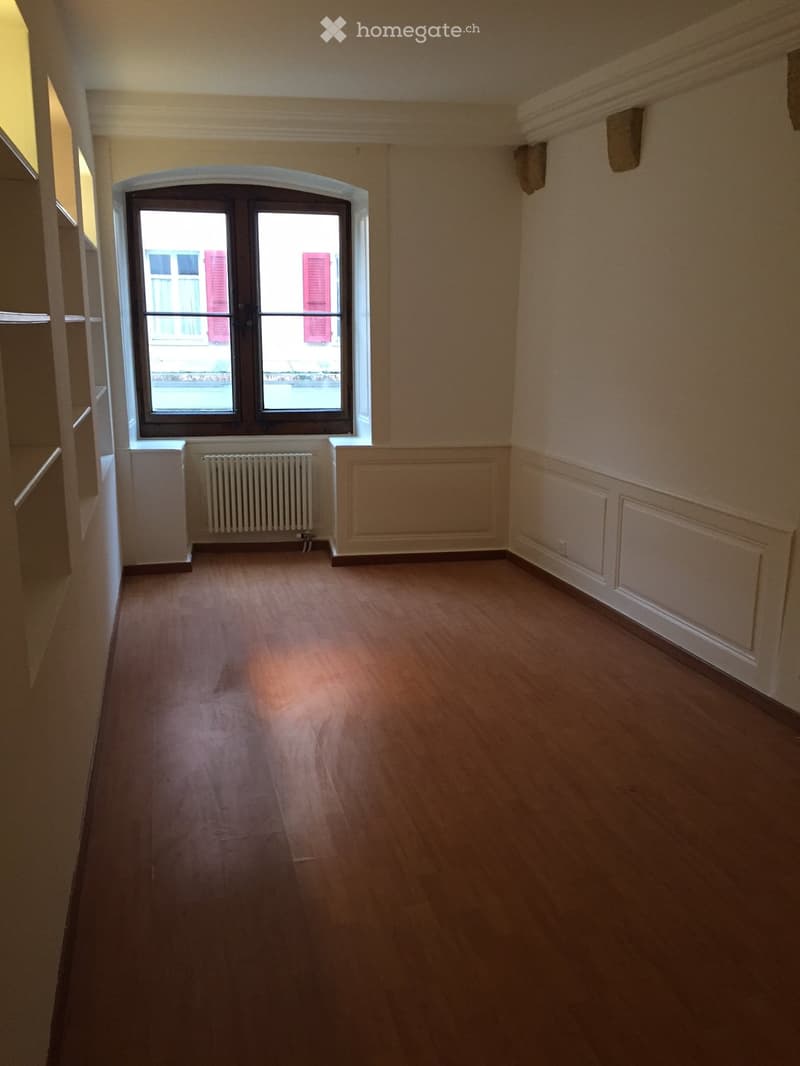 Empty room with wooden flooring, white walls, and a large window with double doors leading outside. A radiator is visible below the window.