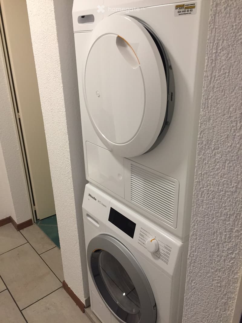 A compact, stacked washing machine and dryer unit in a hallway, with tiled floor and white walls.