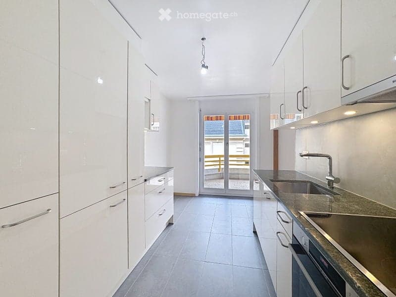 Modern, minimalist kitchen with white cabinets, granite countertop, stainless steel sink and faucet, and built-in oven. Features large glass doors leading to a balcony.