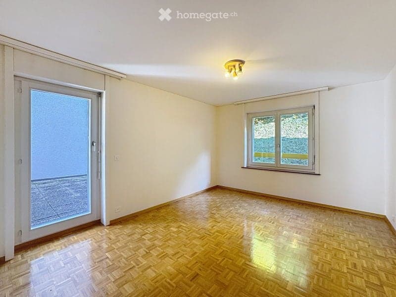 Empty room with wooden flooring, white walls, large window, and sliding glass door leading to a balcony.