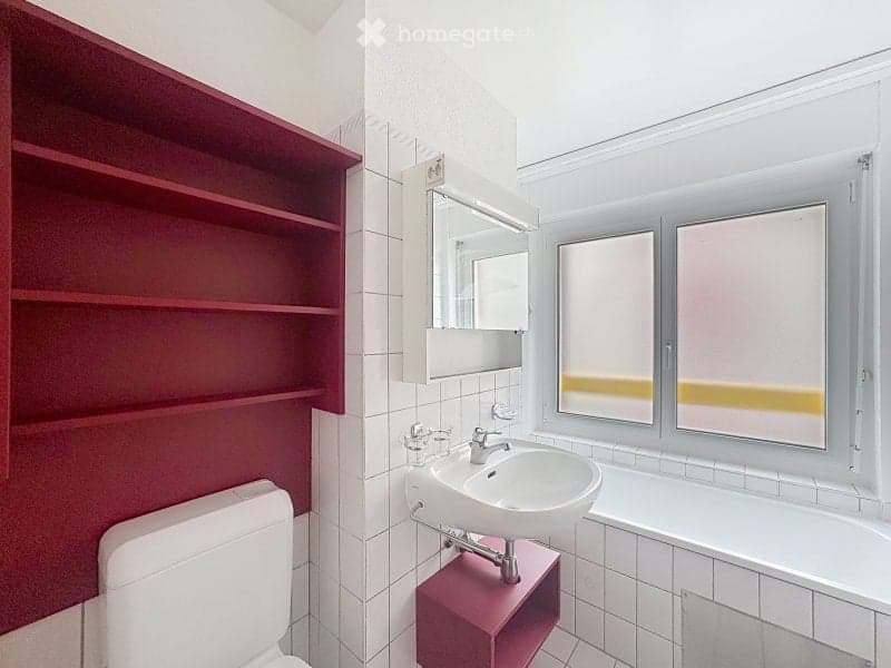 Modern bathroom with white tiles, red shelves, a toilet, a sink with a mirror, and a bathtub. The room features large windows with blinds.