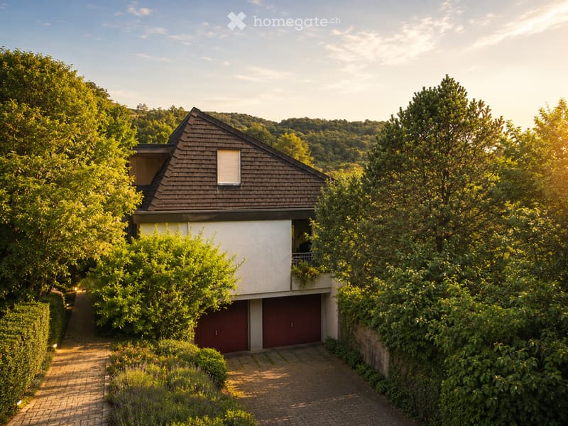 Modern two-story house with a sloped roof, white walls, and a red garage door, surrounded by lush greenery and trees.