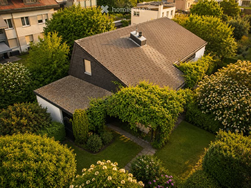 Aerial view of a residential building with a tiled roof, surrounded by lush greenery and flowering bushes.