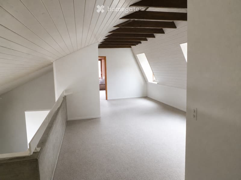 Empty attic space with white walls, carpeted floor, and exposed wooden beams on the ceiling. Features a small window and an open doorway leading to another room.