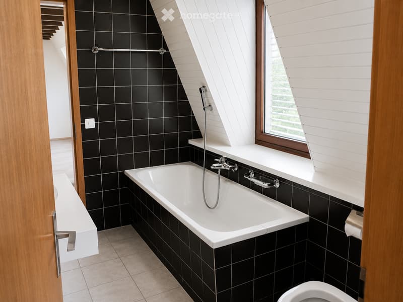 Modern bathroom with black tiled walls, a white bathtub, a toilet, a sink, and a shower hose. The room features a skylight window and wooden trim.