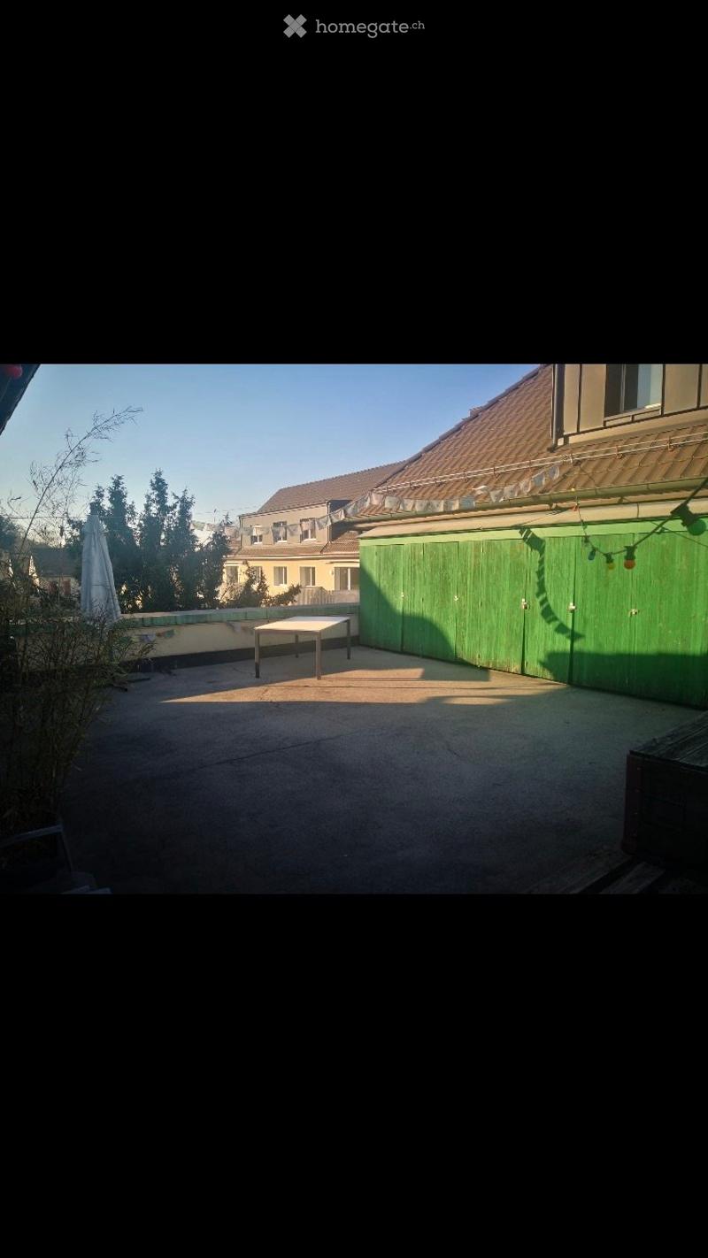 A small outdoor patio area with a concrete floor, a single table, and a green wooden fence. The area is surrounded by trees and neighboring buildings.