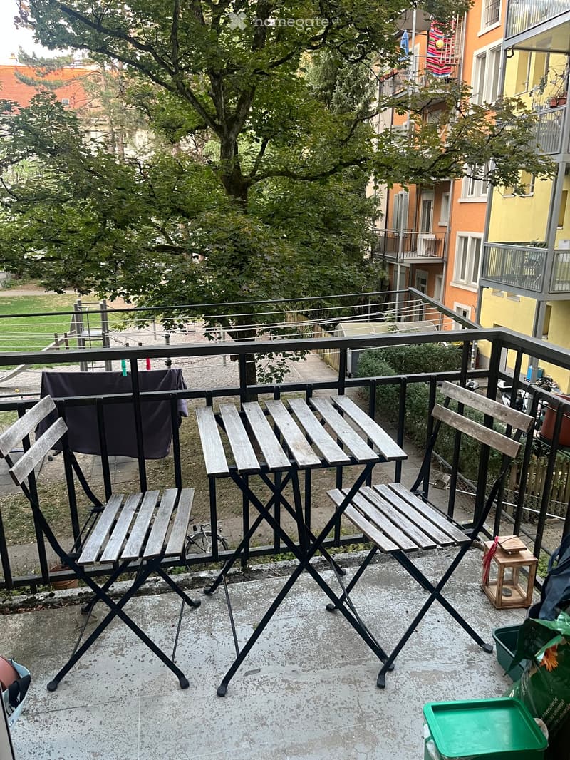 Balcony with wooden folding chairs and a small table, overlooking a residential area with trees and neighboring buildings.
