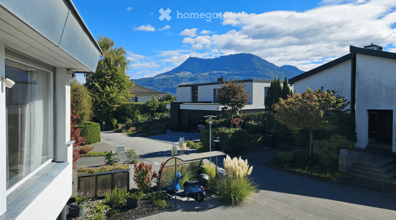 Modern residential complex with clean lines, white facades, and landscaped gardens. A scooter is parked near the entrance, and the background features a scenic mountain view.