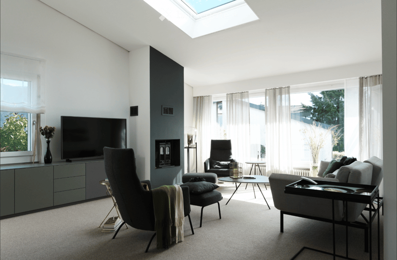 Modern living room with large windows, skylight, grey sofa, coffee table, armchairs, ottoman, TV, floor lamp, and decorative items.
