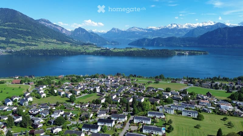 Aerial view of a small village nestled in a valley surrounded by mountains and a lake. The village consists of numerous houses and buildings, with green fields and trees interspersed throughout.