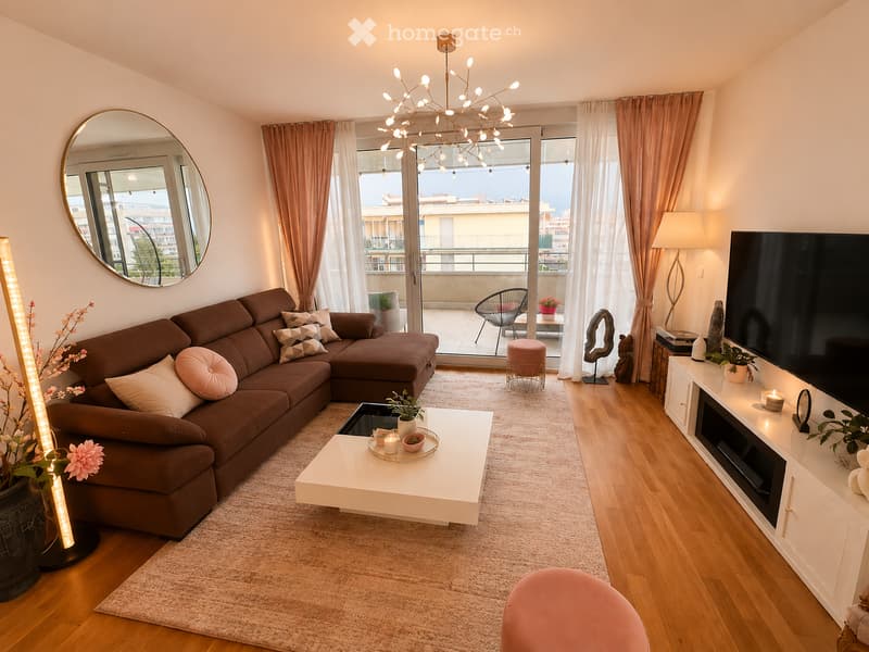 Modern living room with a brown sectional sofa, white coffee table, flat-screen TV on a white stand, floor lamp, and large windows leading to a balcony.