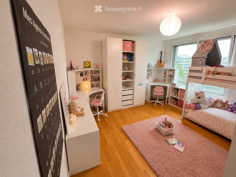 A well-lit children's bedroom with a bunk bed, desk, shelves, and various toys. The room features a large alphabet wall, a pink rug, and a guitar leaning against the wall.
