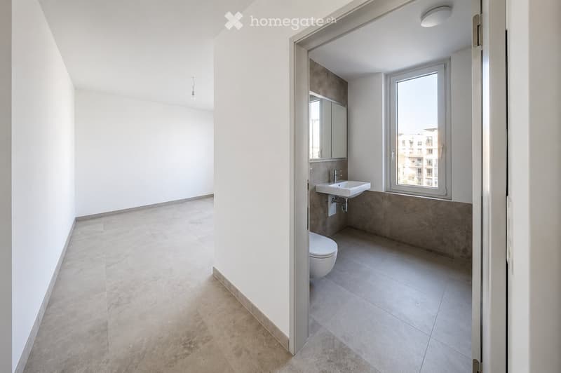 Modern, minimalist bathroom with white walls, beige tiled floor, a sink, toilet, and a mirror. The room is well-lit with natural light from a window showing a cityscape view.