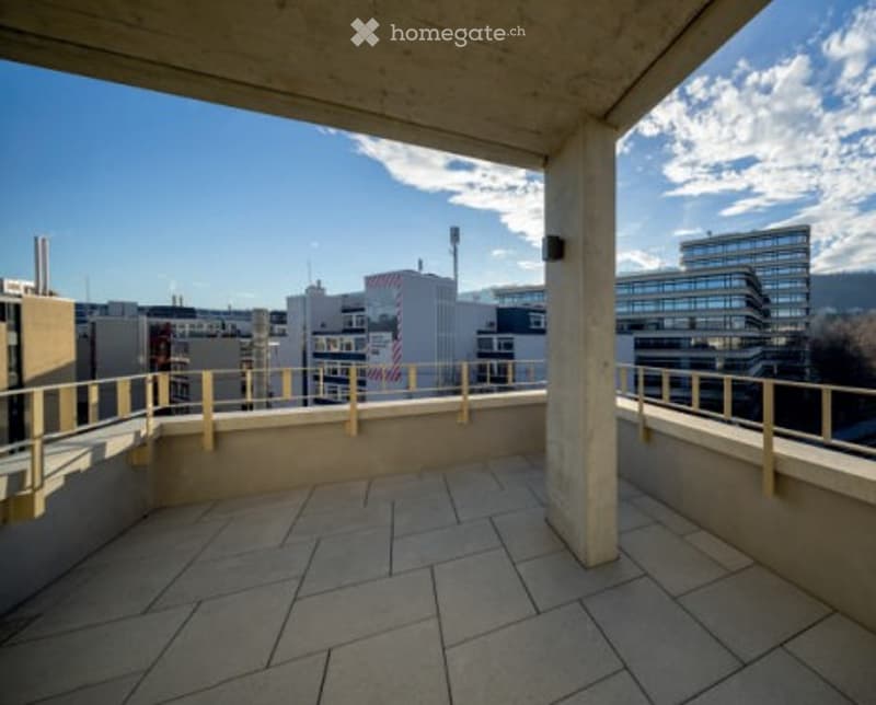 Spacious balcony with tiled floor, metal railings, and a cityscape view of modern buildings and a clear blue sky.