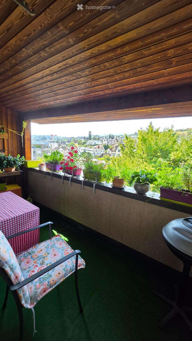 Balcony with wooden ceiling, green artificial turf, a floral-patterned chair, a checkered tablecloth-covered table, and several potted plants on the railing.