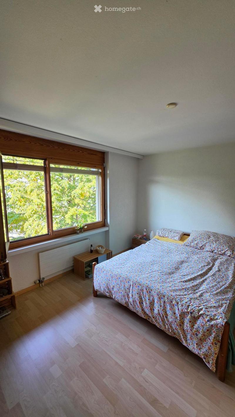 Cozy bedroom with wooden flooring, large window, bed with floral duvet, nightstand, small table, and radiator.