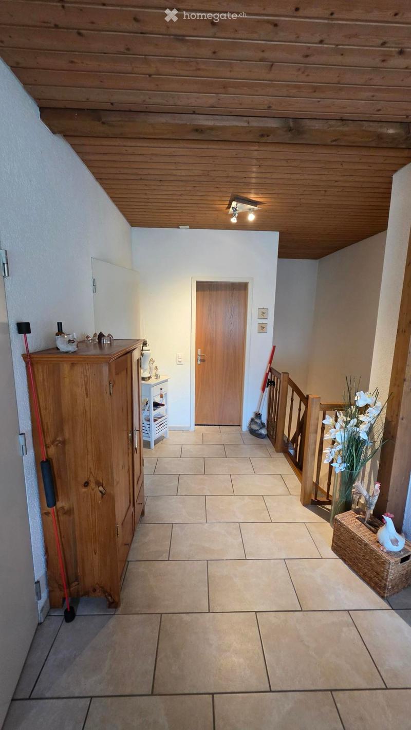Hallway with tiled floor, wooden ceiling, staircase leading upwards, small wooden cabinet, and decorative items including a vase with white flowers and a rooster figurine on a wooden crate.