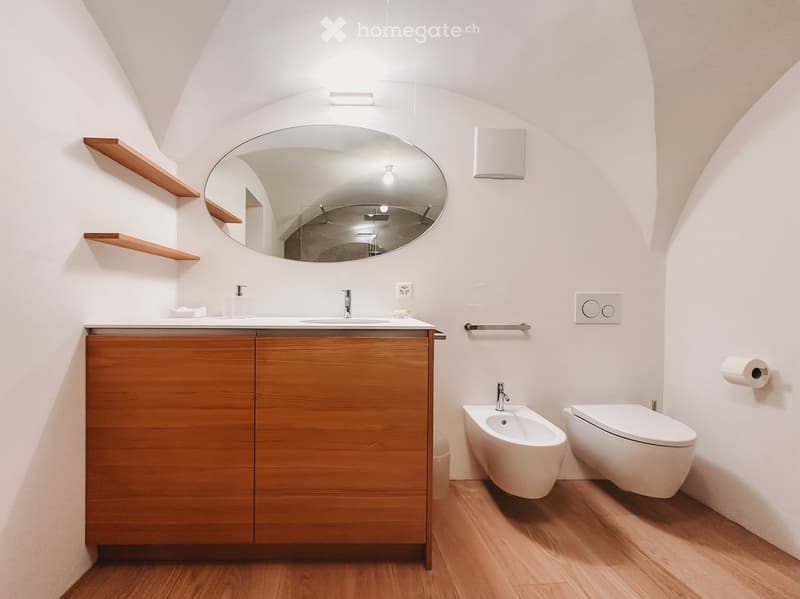 Bathroom. Modern bathroom with wooden floor, white walls, wooden vanity with sink, oval mirror, toilet, bidet, and wooden shelves.