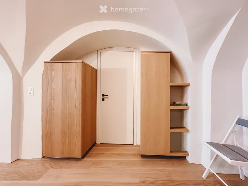 Furnished Room. A small, well-lit room with wooden flooring, white walls, and arched ceilings. Features include a white door, a wooden cabinet, a built-in shelf, and a simple white chair with a grey seat.