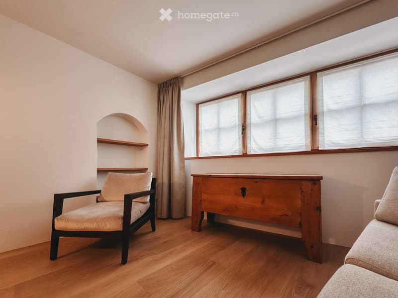 Furnished Room. A modern, minimalist living room with light wooden flooring, large windows with white blinds, a beige sofa, a wooden coffee table, and a single armchair.