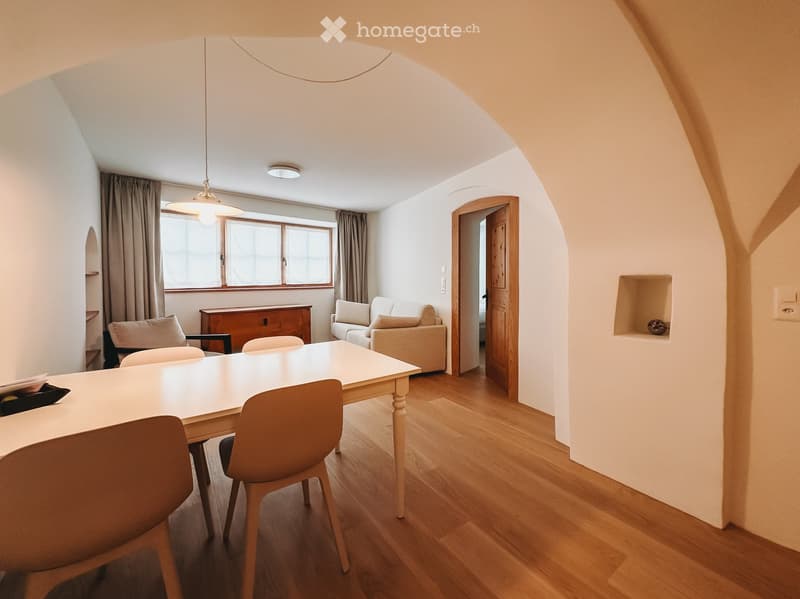 Furnished Room. Bright living and dining area with wooden flooring, large windows, a sofa, dining table with chairs, and a cabinet.