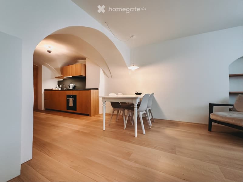 Furnished Room. Open-plan living and dining area with wooden flooring, white dining table, chairs, sofa, and kitchen area with cabinets and appliances.