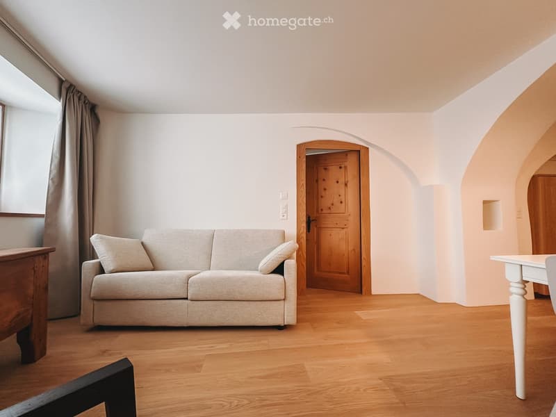 Furnished Room. A cozy living room with a light-colored sofa, wooden floor, and a small table near the window.