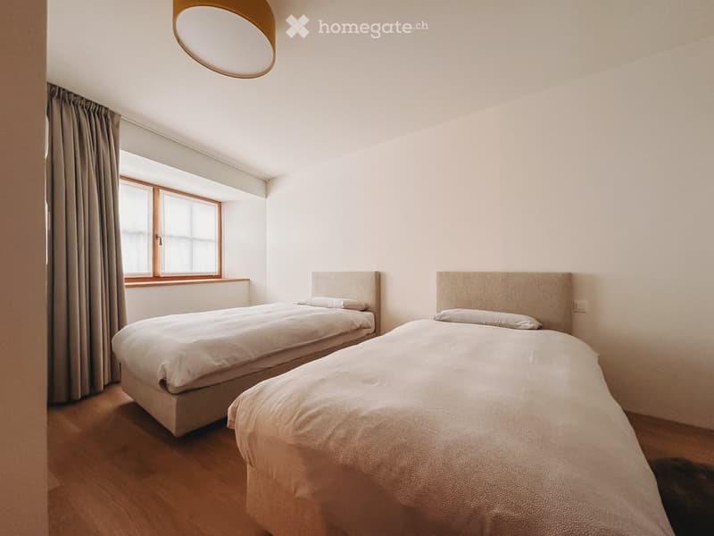 Bedroom. A modern bedroom with two single beds, light-colored bedding, a large window with curtains, and a round wall-mounted lamp.