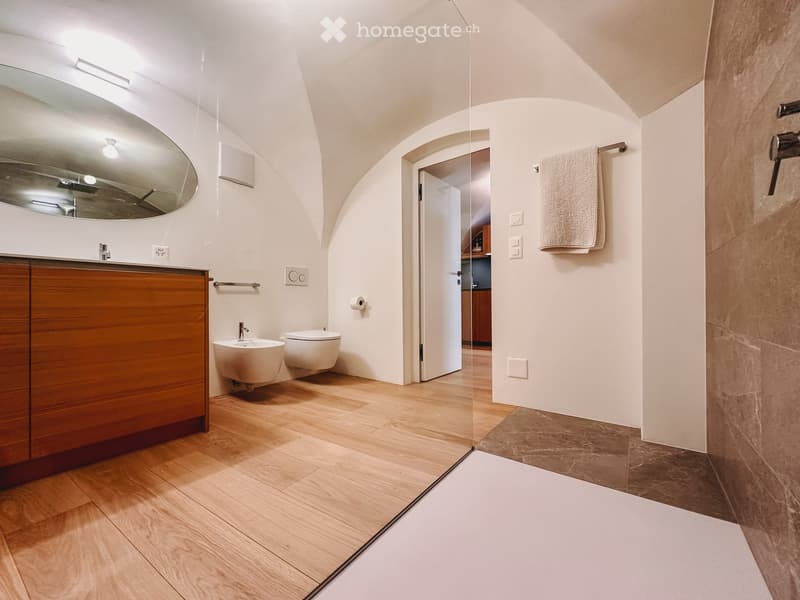 Bathroom. Modern bathroom with wooden flooring, a large oval mirror, a wooden vanity cabinet, a sink, a toilet, and a towel hanging on a rack. The space features a glass partition leading to another room.