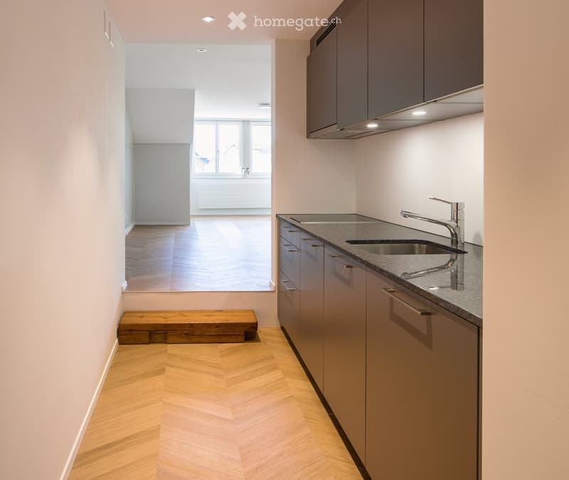 Modern kitchen with grey cabinets, a sink, and a faucet. The floor is herringbone-patterned wood, and there is a wooden bench in the adjacent space.