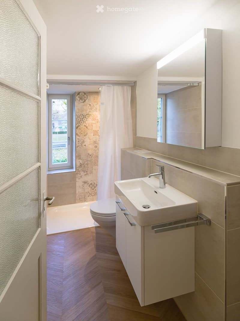 Modern bathroom with a white sink, chrome faucet, large mirror, toilet, and built-in cabinet. Light wood flooring and frosted glass window.