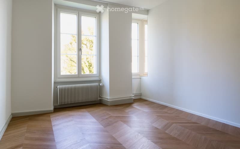 Empty room with wooden herringbone flooring, white walls, and large windows allowing natural light. A radiator is visible under one window.