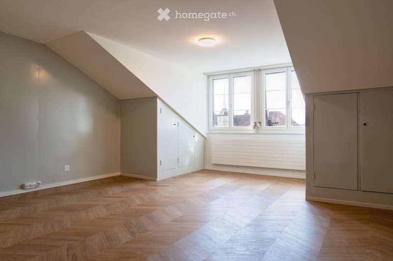 Empty attic room with wooden herringbone flooring, white walls, large windows, built-in closet, and radiator.
