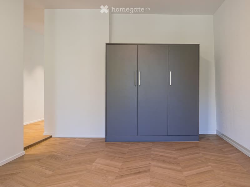 Empty room with light grey walls, wooden herringbone flooring, and a single grey wardrobe with silver handles.