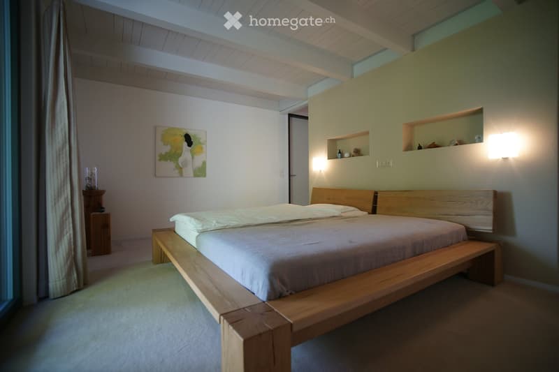 A bedroom with a wooden bed, white walls, and a painting on the wall. The bed has a wooden frame and a mattress with white sheets and pillows. There is a wooden cabinet with a glass vase on top and a curtain on the left side. The wall has three wall niche