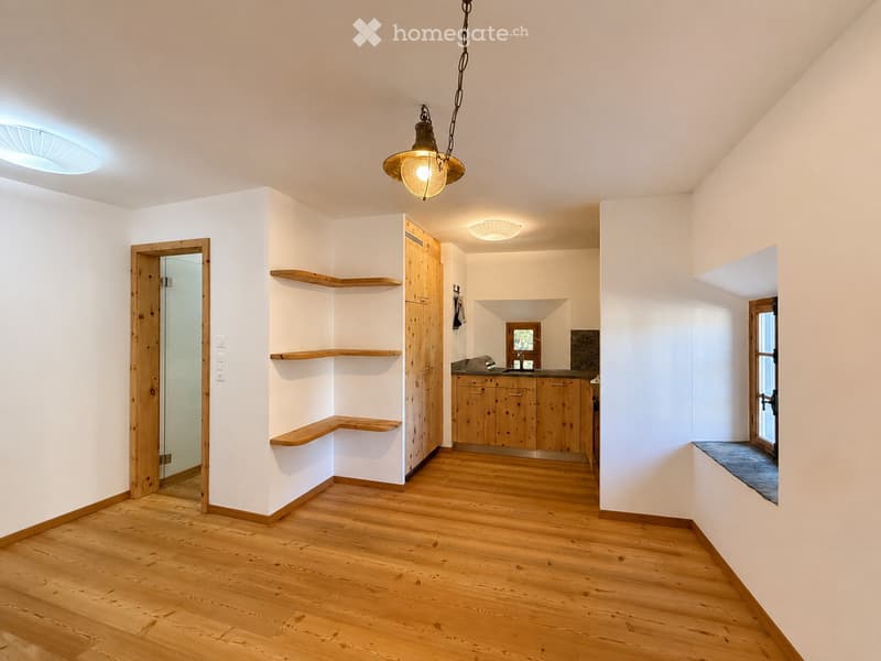 Bright, open space with wooden flooring, white walls, and wooden shelves. A small kitchen area with wooden cabinets and a counter is visible, along with a hanging light fixture and a glass door leading to another room.