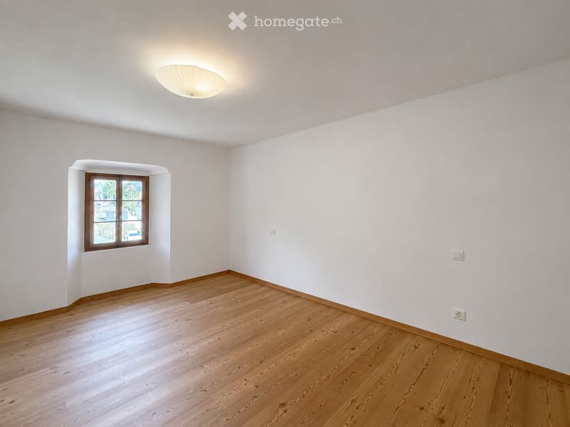 Empty room with light wooden flooring, white walls, and a single window with wooden frame. A modern ceiling light fixture is mounted on the white ceiling.