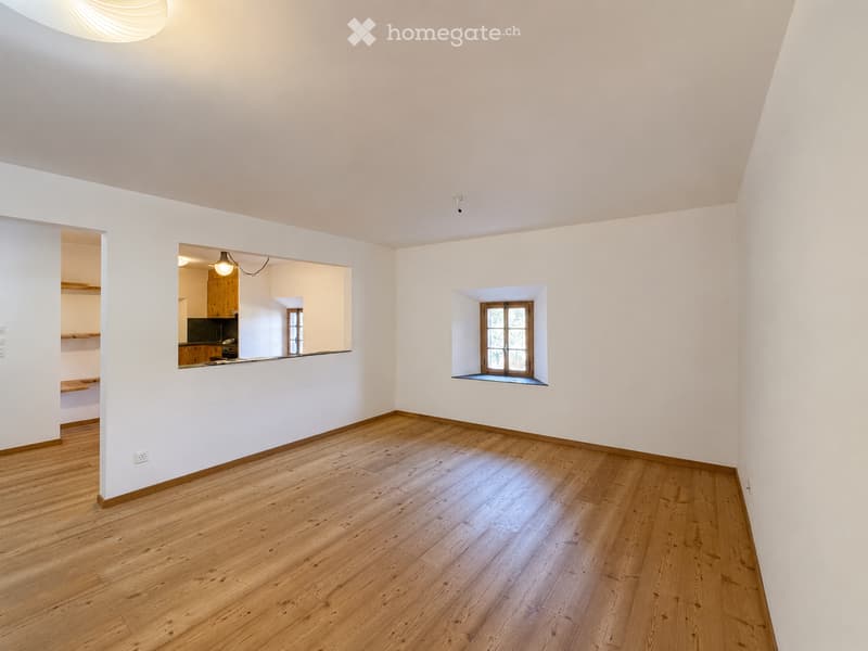 Empty room with wooden flooring and white walls, featuring a small window and an open doorway leading to another room with kitchen elements.